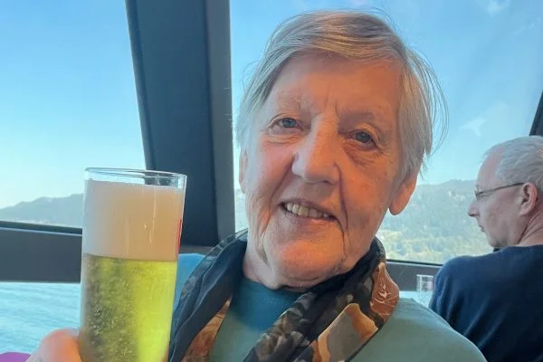 Ältere Frau lächelt am Fenster mit Bergsee im Hintergrund und hält ein Glas Bier mit Schaum.