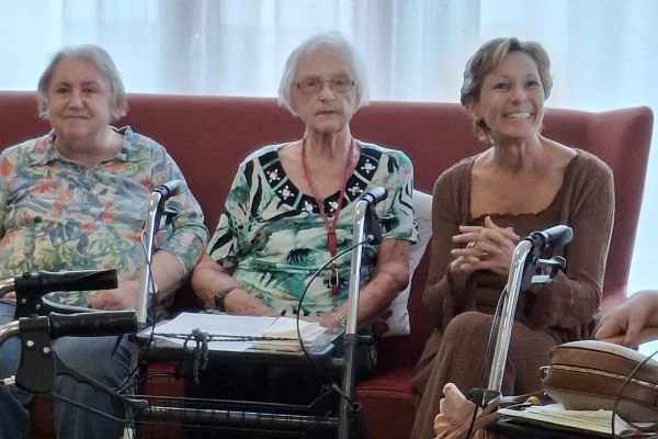 Drei Frauen sitzen auf einem Sofa: zwei ältere Frauen in der Mitte und links mit Rollatoren, rechts eine jüngere Frau, die lächelnd die Hände gefaltet hält.