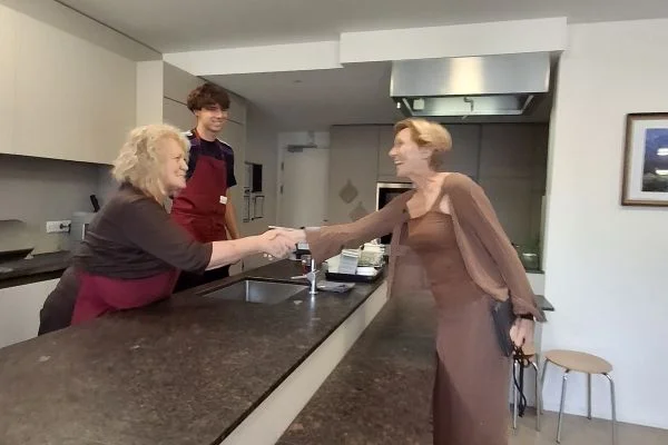 Zwei Frauen schütteln sich lächelnd die Hand über der Küchenarbeitsplatte; ein junger Mann mit Schürze steht hinter der Theke und schaut zu.