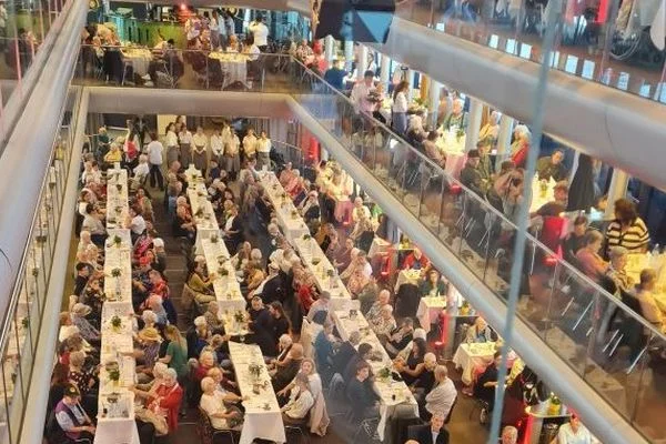 Großer Festsaal mit mehreren langen Tafeln, an denen viele Menschen sitzen und auf zwei Ebenen gemeinsam essen und feiern.