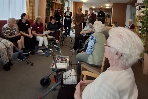 Gruppe älterer Menschen sitzt in einem Gemeinschaftsraum eines Pflegeheims im Stuhlkreis, Pflegekräfte stehen vorn, mehrere Rollatoren sichtbar.