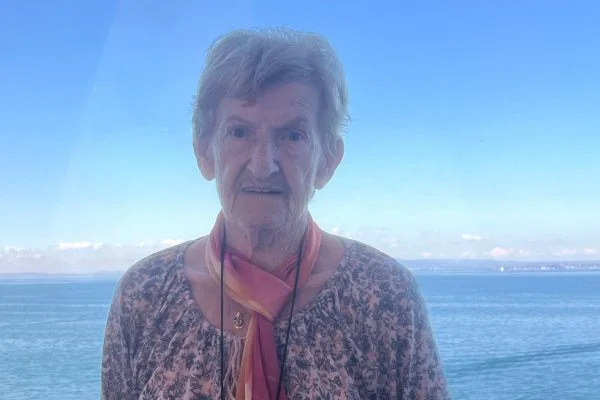 Ältere Frau mit kurzem grauem Haar, gemustertem Oberteil und buntem Schal, blickt in die Kamera; im Hintergrund ruhiges Meer und klarer Himmel
