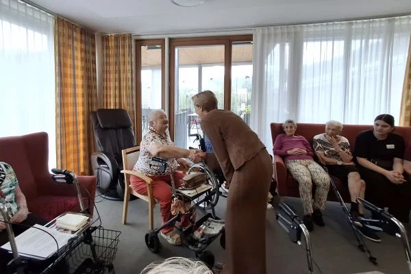 Pflegekraft begrüßt ältere Frau mit Rollator in Gemeinschaftsraum; mehrere Senioren sitzen auf Sofas und Stühlen im hellen Aufenthaltsraum.