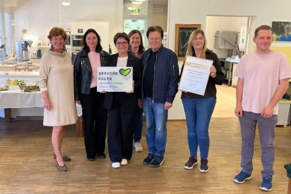 Sieben Menschen stehen in einem Speisesaal vor einer Küche; zwei halten ein Schild mit gesunde Küche und eine Urkunde, alle schauen in die Kamera und lächeln.