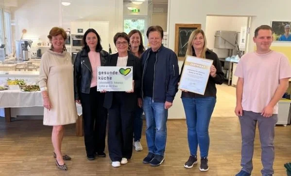 Sieben Menschen stehen in einem Speisesaal vor einer Küche; zwei halten ein Schild mit gesunde Küche und eine Urkunde, alle schauen in die Kamera und lächeln.