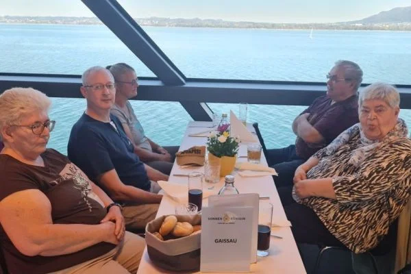 Sechs Erwachsene sitzen an einem langen Tisch in einem Panoramarestaurant auf einem Schiff mit Blick auf einen See; auf dem Tisch Brötchen, Getränke, eine kleine Blumenvase und ein Schild mit der Aufschrift GAISSAU