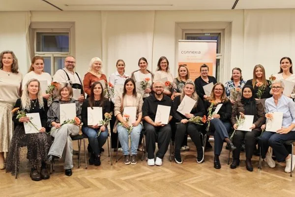 Gruppenfoto von etwa 20 Personen in einem Saal, sitzend und stehend, die Zertifikate und Blumen halten und lächeln bei einer Auszeichnungs- oder Abschlussfeier.