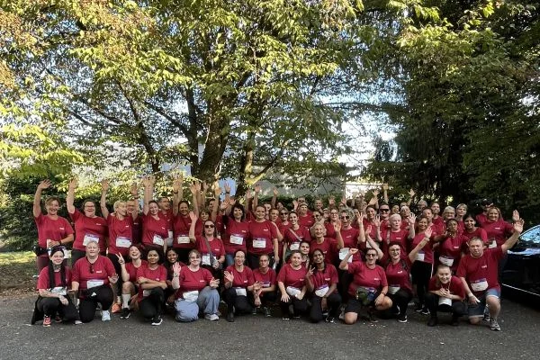 Große Gruppe von etwa 60 Personen in roten T‑Shirts steht unter Bäumen, lächelt und winkt in die Kamera – Teamfoto bei einer Outdoor‑Veranstaltung.