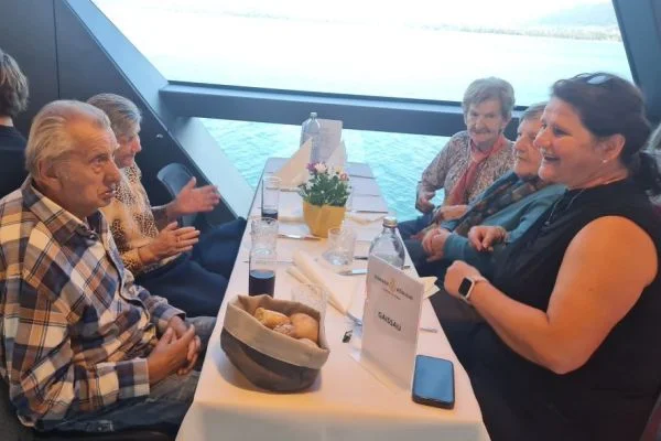 Mehrere ältere Personen sitzen an einem gedeckten Tisch neben großen Fenstern mit Blick aufs Wasser, unterhalten sich und lächeln; auf dem Tisch Brotkorb, Getränke und Gläser.