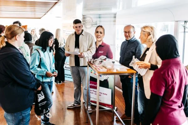 Menschen versammelt um einen kleinen Infostand im Innenraum; Mitarbeitende hinter einem Tisch überreichen Informationsmaterialien und sprechen mit Interessierten.