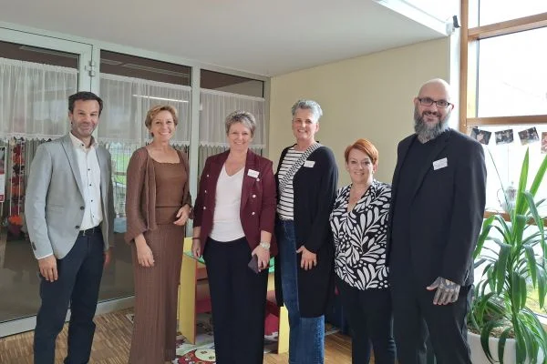 Sechs Erwachsene stehen nebeneinander in einem hellen Raum, lächelnd und formell bis halbformell gekleidet, offenbar bei einer Veranstaltung oder Begrüßung.