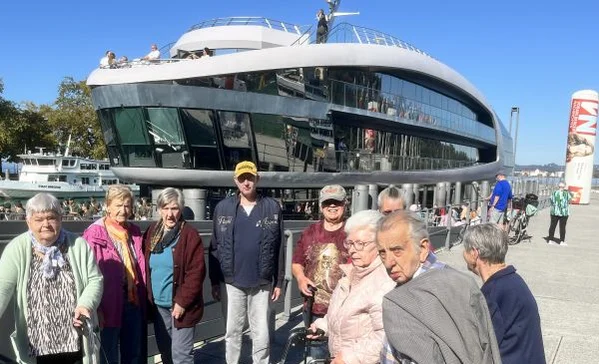 Ausflugsgruppe älterer Menschen steht an einem sonnigen Ufer vor einem modernen, gläsernen Schiffsbau; einige nutzen Rollatoren, andere posieren für ein Foto.