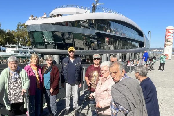 Ausflugsgruppe älterer Menschen steht an einem sonnigen Ufer vor einem modernen, gläsernen Schiffsbau; einige nutzen Rollatoren, andere posieren für ein Foto.