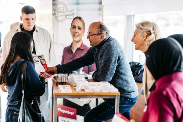 Mehrere Menschen an einem Informationsstand: ein Mann reicht einer jungen Frau einen Flyer, eine lächelnde Frau und weitere Besucher stehen am Tresen mit ausgelegten Informationsmaterialien.